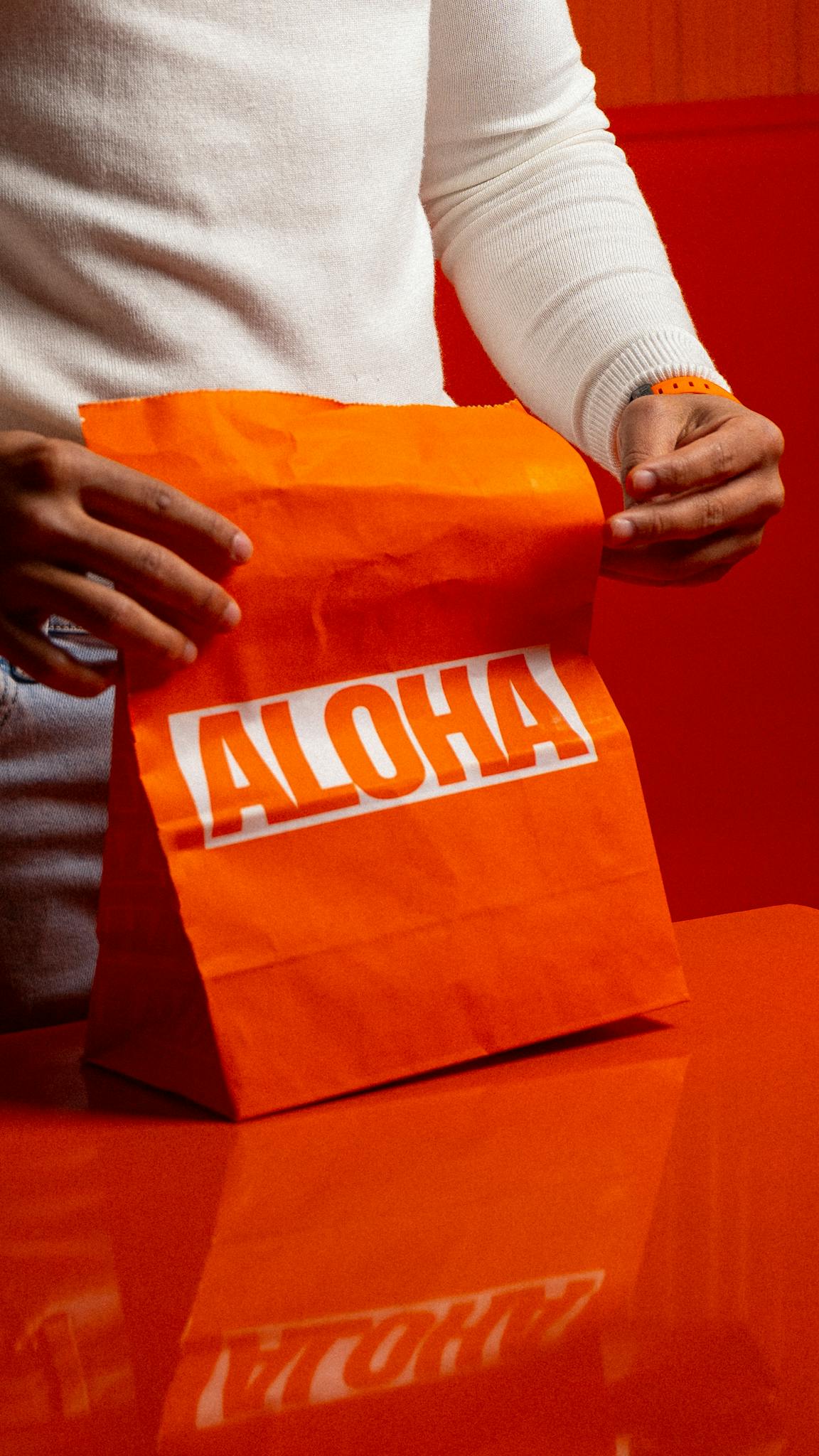 Close-up of hands holding a bright orange 'ALOHA' paper bag on an orange surface. Indoor setting.